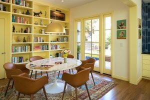A dining area with a round marble table, six brown chairs, and a large pendant light. Bookshelves, wall art, and a rug decorate the yellow-walled room with glass doors leading outside.