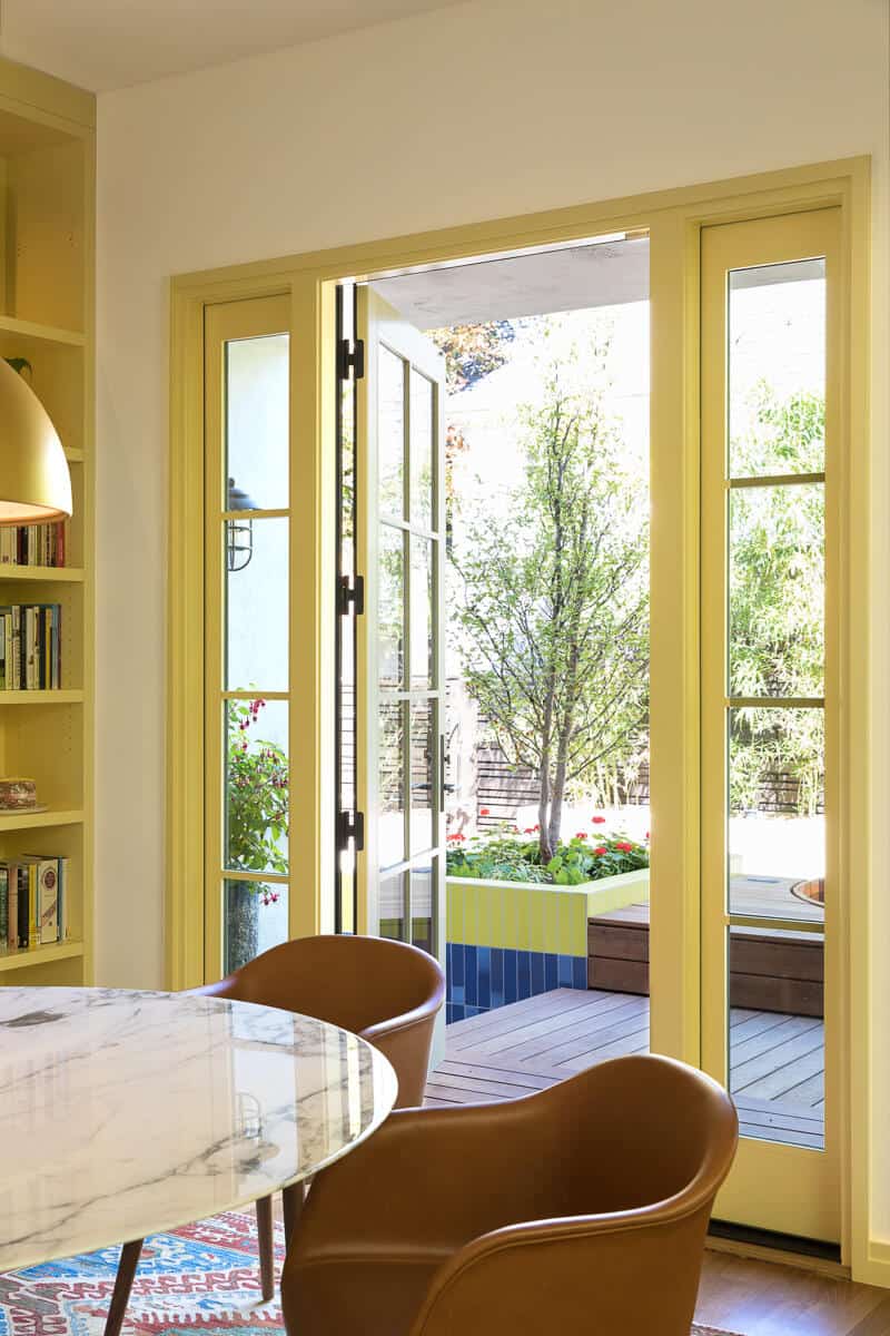 Open yellow-framed glass doors lead from an indoor dining area with a marble table and brown chairs to an outdoor deck with a planter and greenery. Open yellow-framed glass doors lead from an indoor dining area with a marble table and brown chairs to an outdoor deck with a planter and greenery.