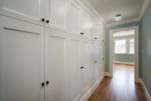 A hallway with light green walls, white ceiling, and built-in white cabinets running along one side. The hardwood floor leads to a room with a window at the far end.