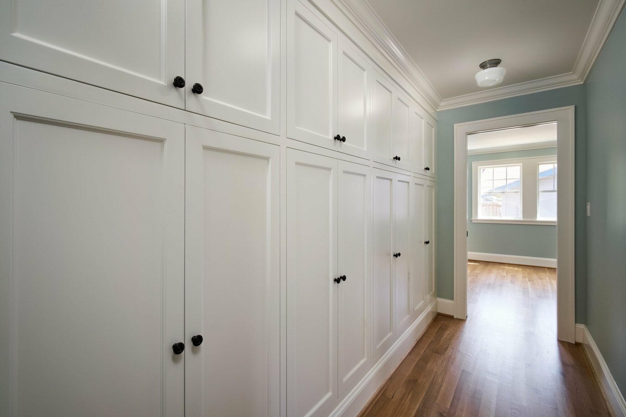 A hallway with light green walls, white ceiling, and built-in white cabinets running along one side. The hardwood floor leads to a room with a window at the far end. A hallway with light green walls, white ceiling, and built-in white cabinets running along one side. The hardwood floor leads to a room with a window at the far end.