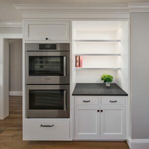A kitchen with a built-in stainless steel double oven, adjacent open white shelving with a small plant and books, and white cabinets with black handles against a light gray wall and wooden floor.