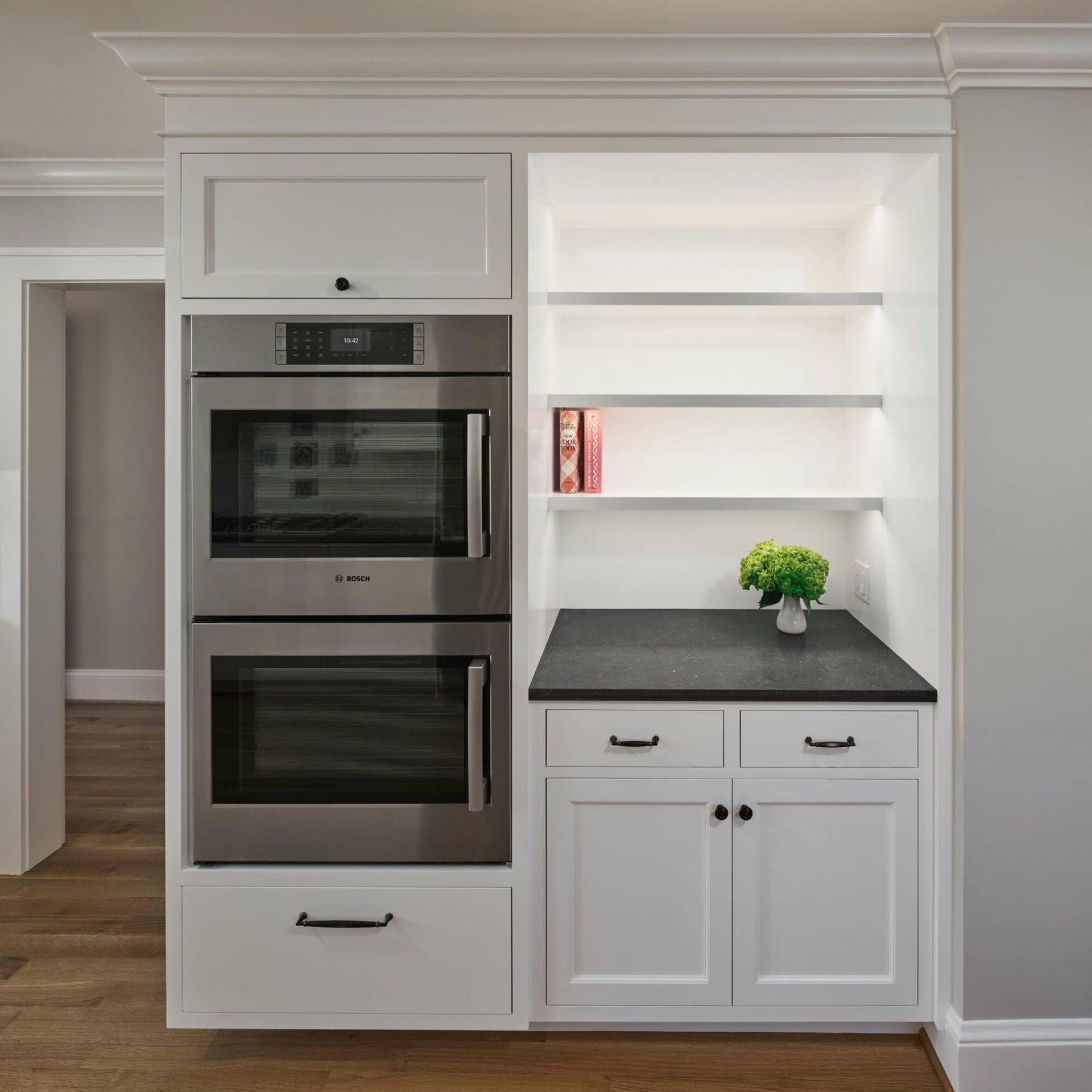 A kitchen with a built-in stainless steel double oven, adjacent open white shelving with a small plant and books, and white cabinets with black handles against a light gray wall and wooden floor. A kitchen with a built-in stainless steel double oven, adjacent open white shelving with a small plant and books, and white cabinets with black handles against a light gray wall and wooden floor.