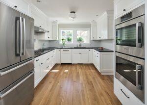 Modern kitchen with white cabinets, stainless steel appliances, and wooden flooring. Natural light enters through a window above the sink.