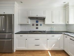 A modern kitchen with white cabinets, black countertops, stainless steel appliances, a gas stove, and decorative tiles above the stove, illuminated by natural light.