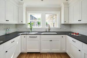 A modern kitchen with white cabinets, black countertops, a center sink beneath a double window, and light wood flooring. Items on the counter include a vase of flowers, bottled beverages, and apples.