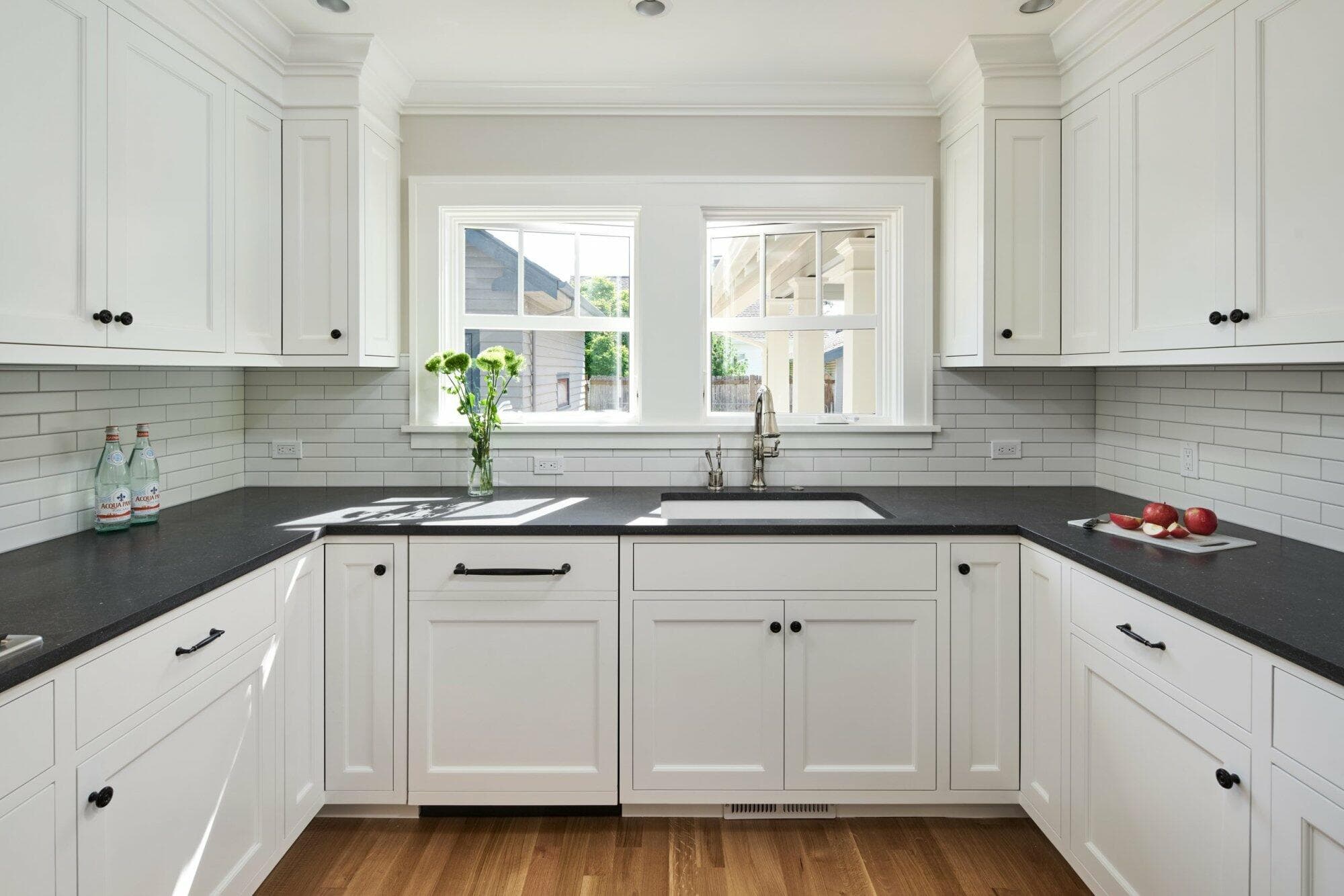 A modern kitchen with white cabinets, black countertops, a center sink beneath a double window, and light wood flooring. Items on the counter include a vase of flowers, bottled beverages, and apples. A modern kitchen with white cabinets, black countertops, a center sink beneath a double window, and light wood flooring. Items on the counter include a vase of flowers, bottled beverages, and apples.