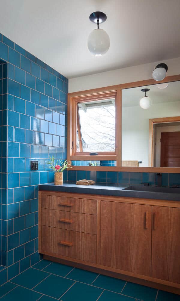 Bathroom with blue tiled walls and floor, a wooden vanity with black countertop and drawers, large mirror, two round light fixtures, and a small window.