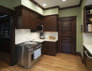 A kitchen with wooden cabinetry, a stainless steel gas stove, a microwave, and a countertop with kitchen appliances and storage jars. The room has hardwood floors and a wooden door.