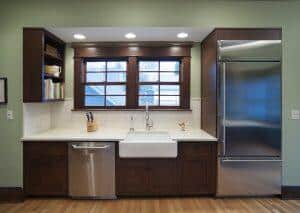 A kitchen with green walls features a large sink under two windows, a stainless steel dishwasher, a knife block, a refrigerator, and open shelves with books above the countertop.