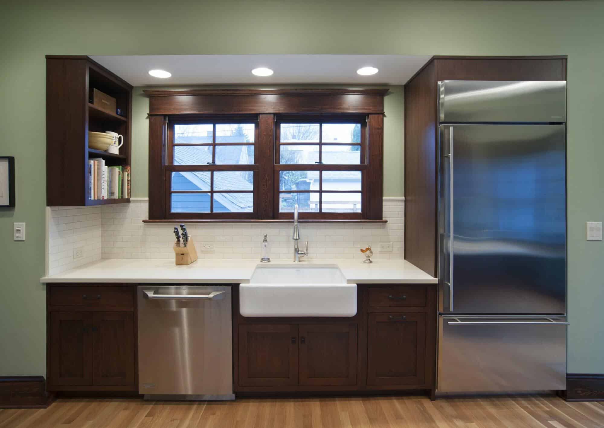 A kitchen with green walls features a large sink under two windows, a stainless steel dishwasher, a knife block, a refrigerator, and open shelves with books above the countertop. A kitchen with green walls features a large sink under two windows, a stainless steel dishwasher, a knife block, a refrigerator, and open shelves with books above the countertop.