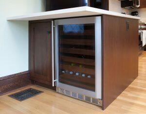 Built-in wine cooler with a glass door and stainless steel frame, situated under a countertop in a kitchen with wood cabinets and hardwood flooring.