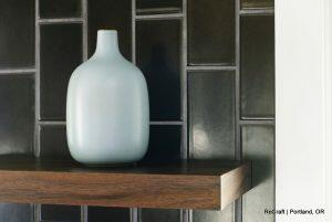 A light blue vase sits on a wooden shelf against a dark tiled wall. Text in the bottom right corner reads 