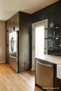 A modern kitchen with dark wood cabinets, stainless steel refrigerator and dishwasher, a door leading outside, black tile backsplash, and wooden flooring.