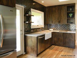 Modern kitchen featuring dark wood cabinets, stainless steel appliances, and white countertops. Black tile backsplash and a farmhouse sink with white bowl and small shelves above. Window and door to the left.