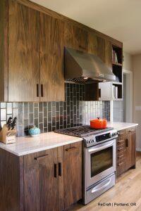 A modern kitchen with dark wooden cabinets, stainless steel stove and hood, white countertops, and a black tiled backsplash. There is a knife block, a blue teapot, and an orange pot on the stove.