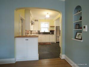 A view of a modern kitchen with white cabinets, stainless steel appliances, and dark tiled flooring, seen through an arched doorway from an adjoining room.