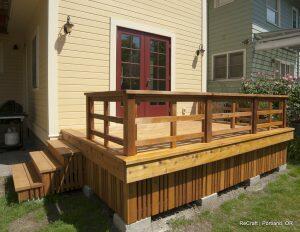 A wooden deck with railings and stairs is attached to the back of a yellow house with red-framed double doors. A green house is partially visible on the right.