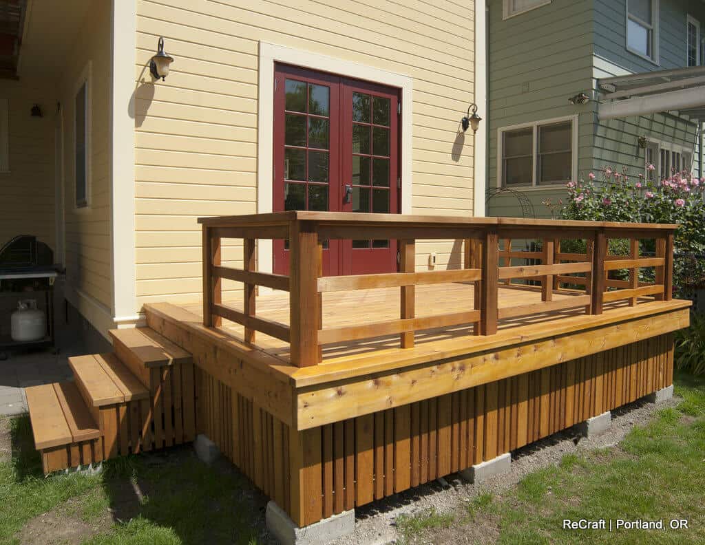 A wooden deck with railings and stairs is attached to the back of a yellow house with red-framed double doors. A green house is partially visible on the right. A wooden deck with railings and stairs is attached to the back of a yellow house with red-framed double doors. A green house is partially visible on the right.