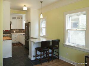 A modern kitchen features light green walls, dark tile flooring, white cabinetry, stainless steel appliances, a hanging light fixture, and a breakfast bar with two chairs.