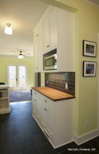 A kitchen with light green walls, black tile flooring, and white cabinets. There is a wooden countertop, a backsplash with dark tiles, and a door leading to a well-lit outdoor area.