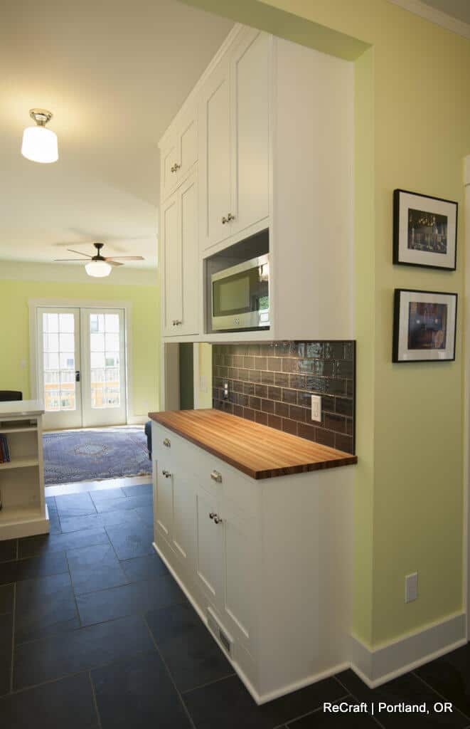 A kitchen with light green walls, black tile flooring, and white cabinets. There is a wooden countertop, a backsplash with dark tiles, and a door leading to a well-lit outdoor area. A kitchen with light green walls, black tile flooring, and white cabinets. There is a wooden countertop, a backsplash with dark tiles, and a door leading to a well-lit outdoor area.