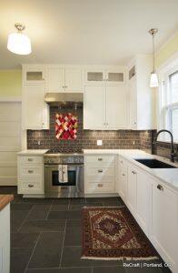A modern kitchen with white cabinetry, a stainless steel stove and range hood, black tiled backsplash, a black sink, and a red patterned rug on the dark tiled floor.