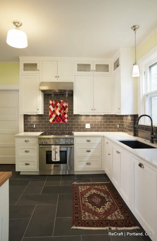 A modern kitchen with white cabinetry, a stainless steel stove and range hood, black tiled backsplash, a black sink, and a red patterned rug on the dark tiled floor. A modern kitchen with white cabinetry, a stainless steel stove and range hood, black tiled backsplash, a black sink, and a red patterned rug on the dark tiled floor.