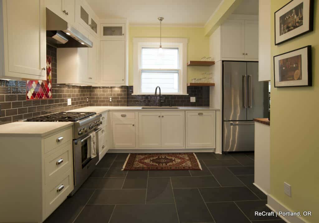 A modern kitchen with white cabinets, stainless steel appliances, a black subway tile backsplash, slate flooring, and a patterned rug near the sink. A window above the sink allows natural light in. A modern kitchen with white cabinets, stainless steel appliances, a black subway tile backsplash, slate flooring, and a patterned rug near the sink. A window above the sink allows natural light in.
