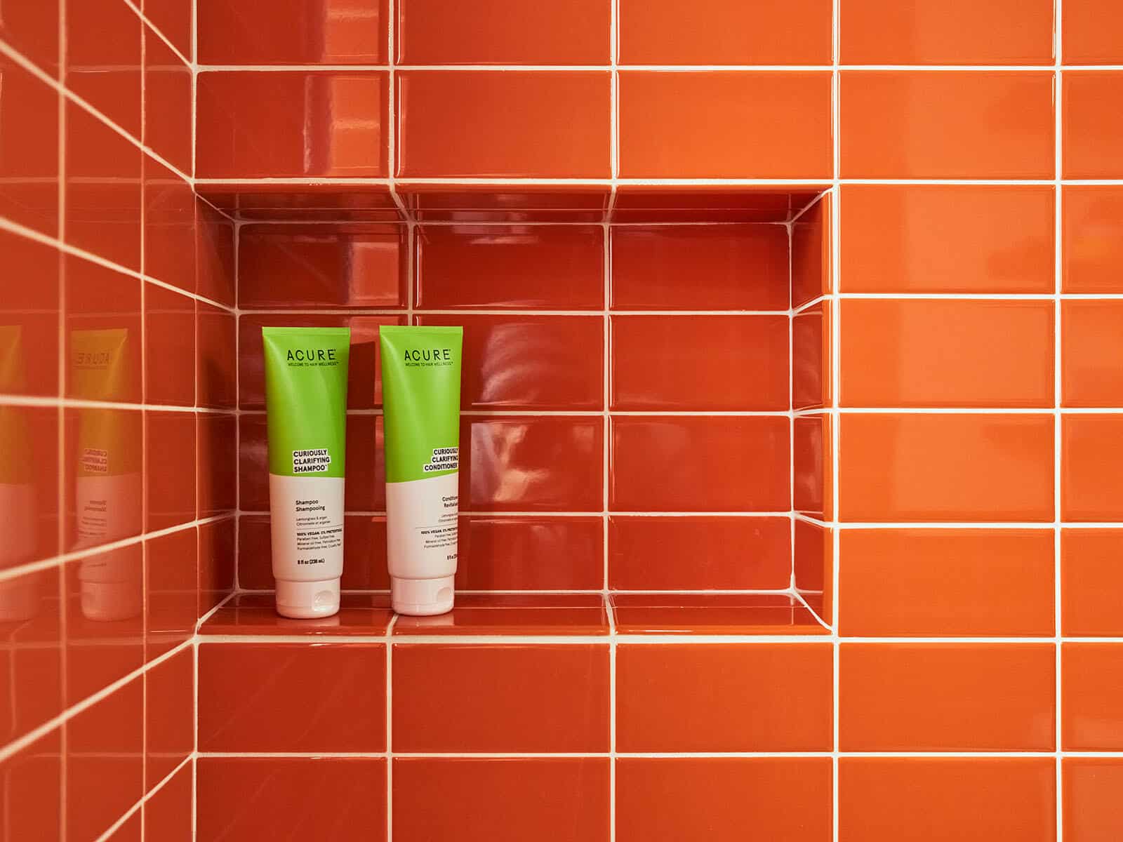 Two green and white Acure shampoo and conditioner bottles are placed on a shelf in an orange tiled shower.