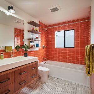 A modern bathroom featuring a white bathtub with an orange-tiled wall, a white sink, wooden cabinets, hexagonal floor tiles, and floating shelves adorned with decor items.