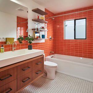 A modern bathroom with orange tiled walls, a white bathtub, a vanity with a sink and mirror, floating shelves, a window, and a floor with hexagonal white tiles.