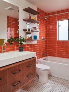 A modern bathroom with bright orange subway tiles, white fixtures, a wooden vanity with a sink, and open shelves displaying decorative items above the toilet.