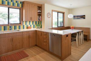 A modern kitchen with light wood cabinetry, white countertops, colorful striped backsplash, stainless steel dishwasher, sink by a window, and a dining nook with a bench and chairs by another window.