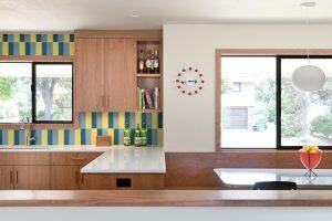 A modern kitchen with wooden cabinets, colorful tiled backsplash, a sink by the window, and a built-in counter attached to a dining area with a black chair and a bowl of fruit.