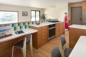 Modern kitchen featuring wooden cabinets, a colorful tiled backsplash, and a built-in oven. A woman in a red dress is walking by, and a tablet is placed on the countertop.
