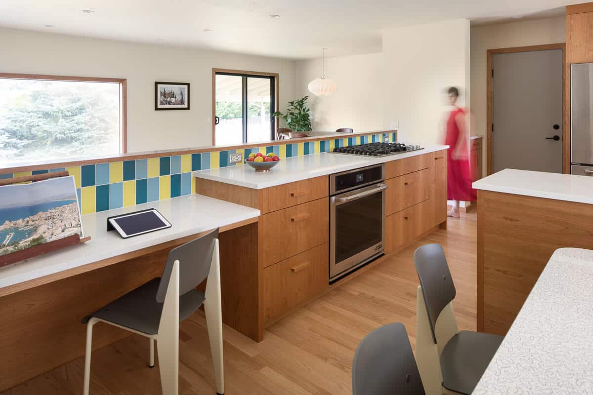 Modern kitchen featuring wooden cabinets, a colorful tiled backsplash, and a built-in oven. A woman in a red dress is walking by, and a tablet is placed on the countertop. Modern kitchen featuring wooden cabinets, a colorful tiled backsplash, and a built-in oven. A woman in a red dress is walking by, and a tablet is placed on the countertop.