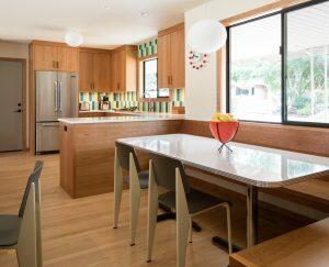 A kitchen with wooden cabinetry, a center island, stainless steel appliances, a dining table with chairs, and a fruit bowl on the table. A large window offers natural light and outdoor view.