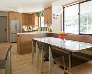 Modern kitchen with wooden cabinets, an island with a stainless steel fridge, a built-in dining nook with a table and chairs, a bowl of apples, and a window letting in natural light.