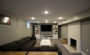 A basement living room with a black sectional sofa, white coffee table, a large TV on a white entertainment center, and a white brick fireplace. The room has grey walls and carpet flooring.