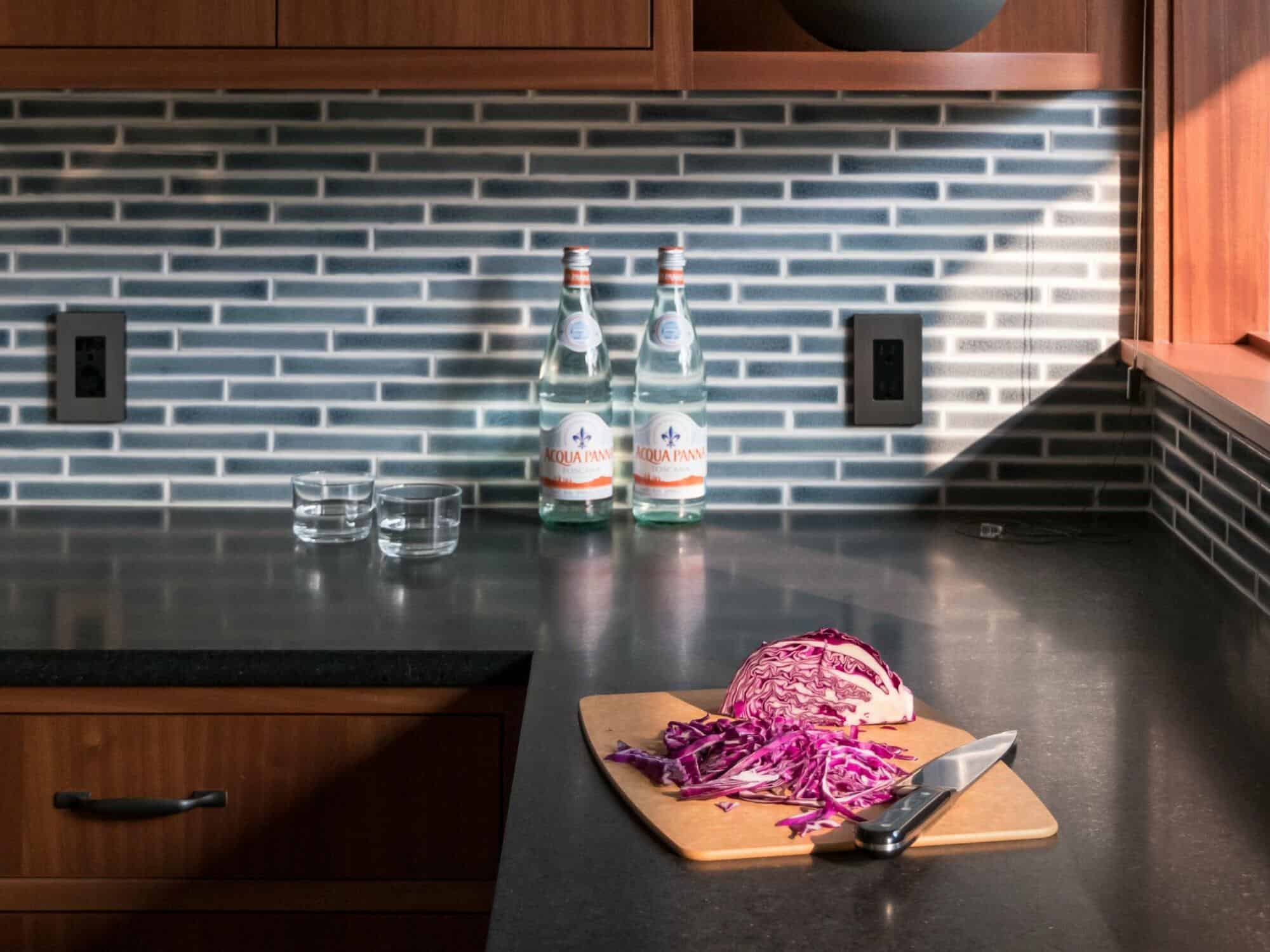 A cutting board with chopped red cabbage and a knife on a kitchen counter. Two bottles of mineral water and two glasses are on the counter against a tile backsplash. A cutting board with chopped red cabbage and a knife on a kitchen counter. Two bottles of mineral water and two glasses are on the counter against a tile backsplash.