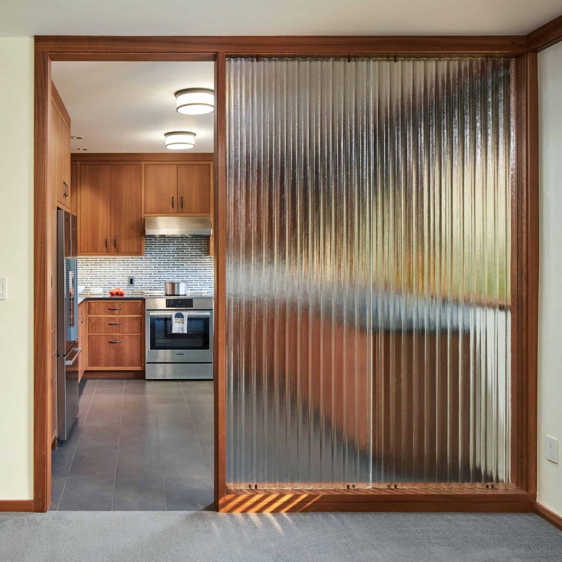 A modern kitchen with wooden cabinets, stainless steel appliances, and a tiled backsplash is visible through a fluted glass partition. A carpeted area is in the foreground. A modern kitchen with wooden cabinets, stainless steel appliances, and a tiled backsplash is visible through a fluted glass partition. A carpeted area is in the foreground.