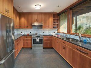 A modern U-shaped kitchen with wooden cabinets, stainless steel appliances, a tiled backsplash, and large windows providing natural light.