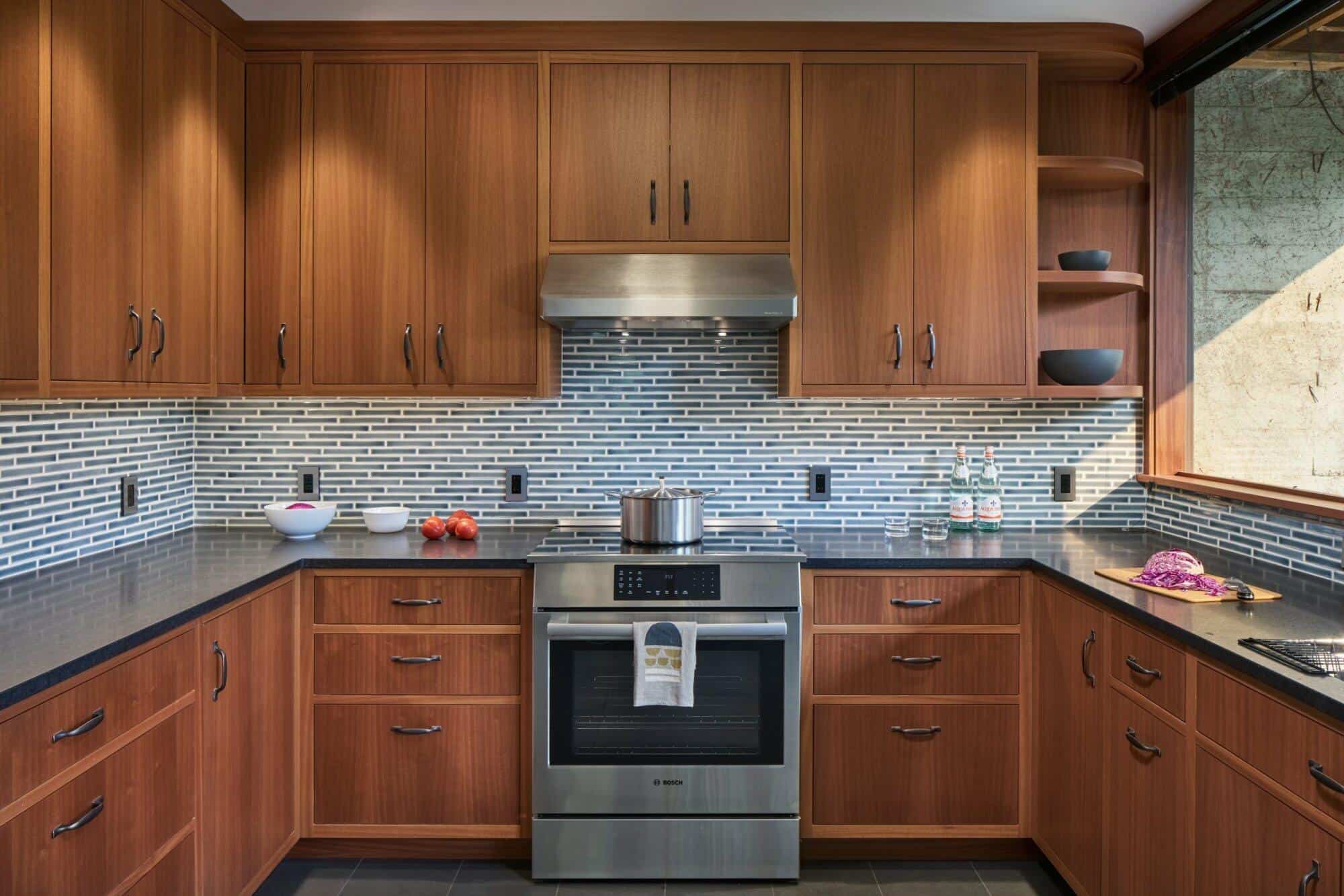Modern kitchen with wood cabinets, stainless steel oven, and a black countertop. Backsplash features horizontal gray and white tiles. A pot is on the stove, and kitchen items are placed around. Modern kitchen with wood cabinets, stainless steel oven, and a black countertop. Backsplash features horizontal gray and white tiles. A pot is on the stove, and kitchen items are placed around.