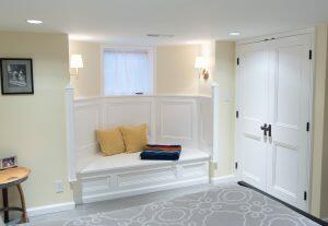A cozy nook with pillows and a blanket, illuminated by wall sconces, is adjacent to a set of double doors and a wooden side table with a picture frame.