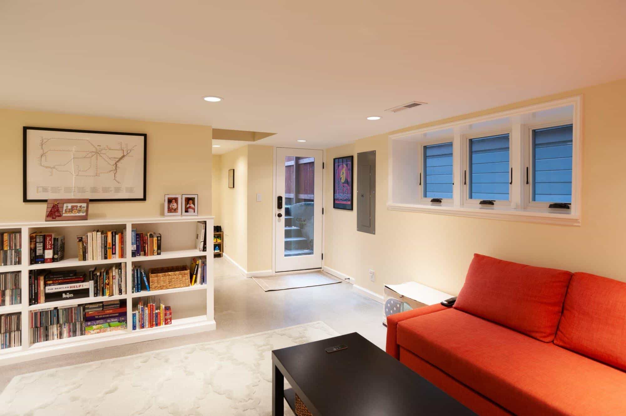 Modern basement living room with orange sofa, white bookshelf filled with books, and natural light from multiple windows, showcasing ReCraft Home Remodeling's design aesthetic.