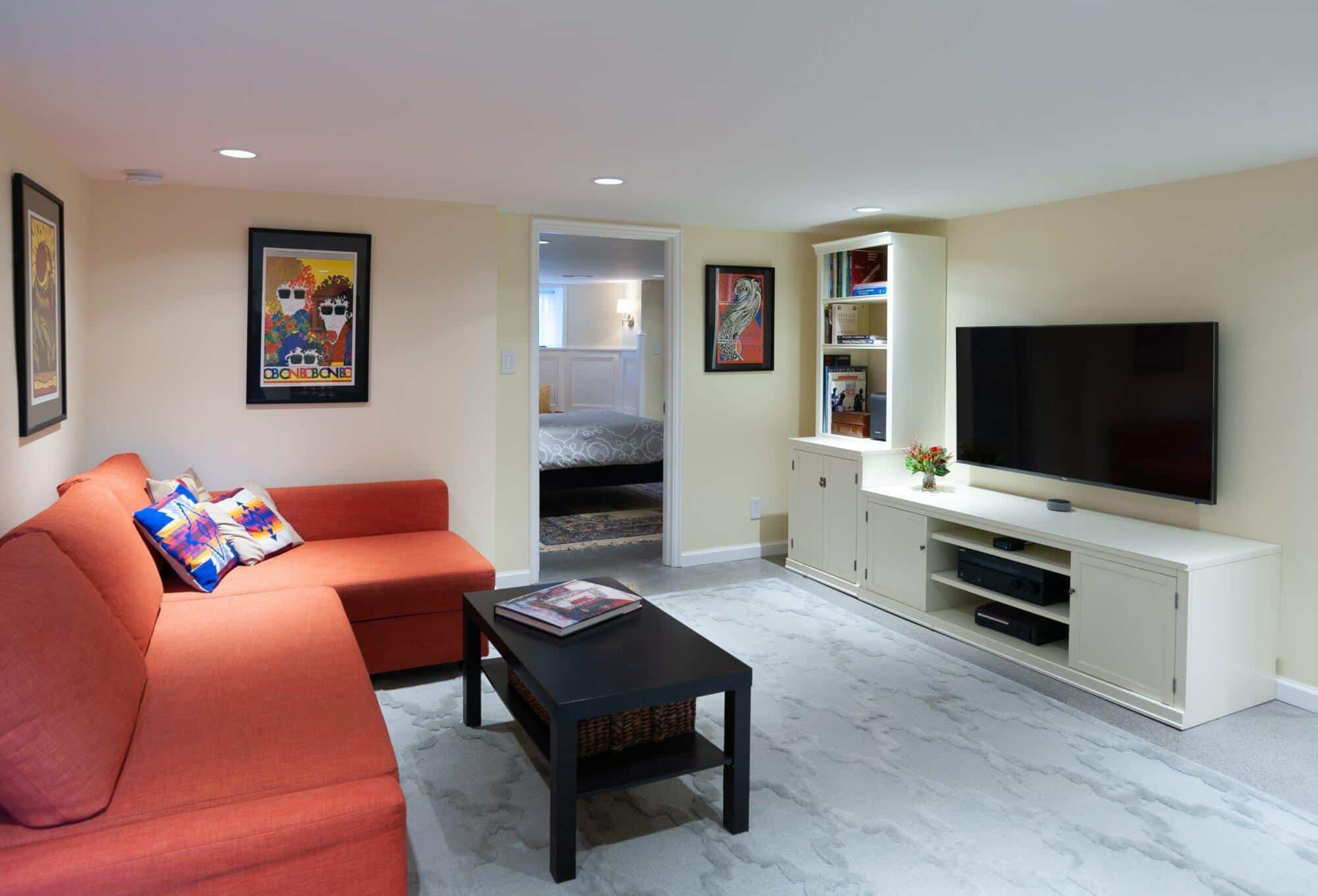 Cozy basement living room featuring an orange sectional sofa, black coffee table, and wall-mounted TV, with decorative artwork and shelves, showcasing ReCraft Home Remodeling's design expertise.