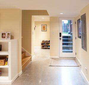 A basement entryway with a white door, stairs to the left, a shoe rack, and framed photos on a shelf. The floor is concrete, and there is a utility panel on the right wall.