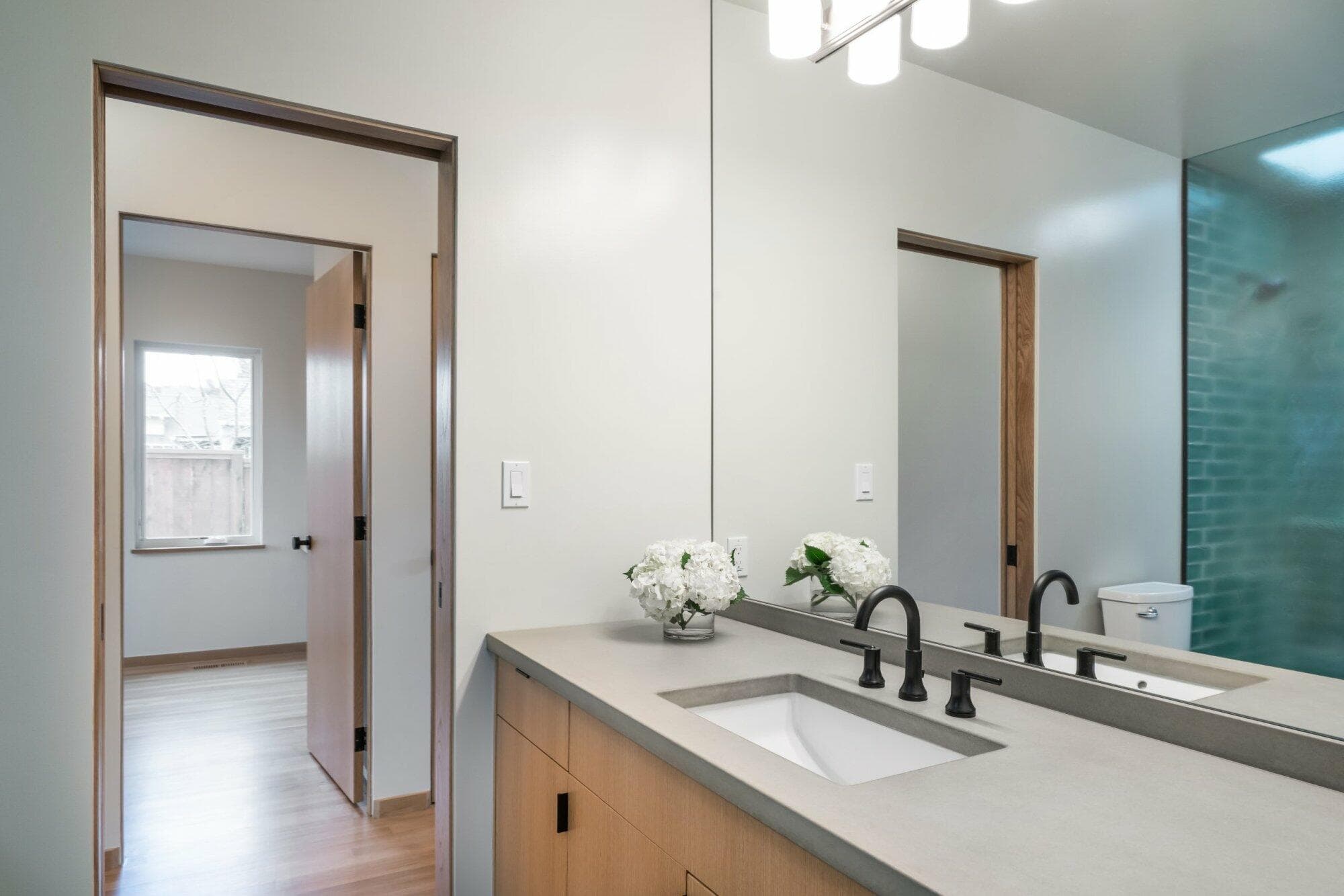 Modern bathroom with a large mirror, double sink, black faucets, light fixtures, and a vase of white flowers. A doorway leads to another room with natural light from a window.