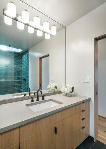 Modern bathroom with a large mirror, lighted vanity fixture, single sink with a black faucet, and a wooden cabinet. The shower area in the background has teal subway tiles and a glass door.