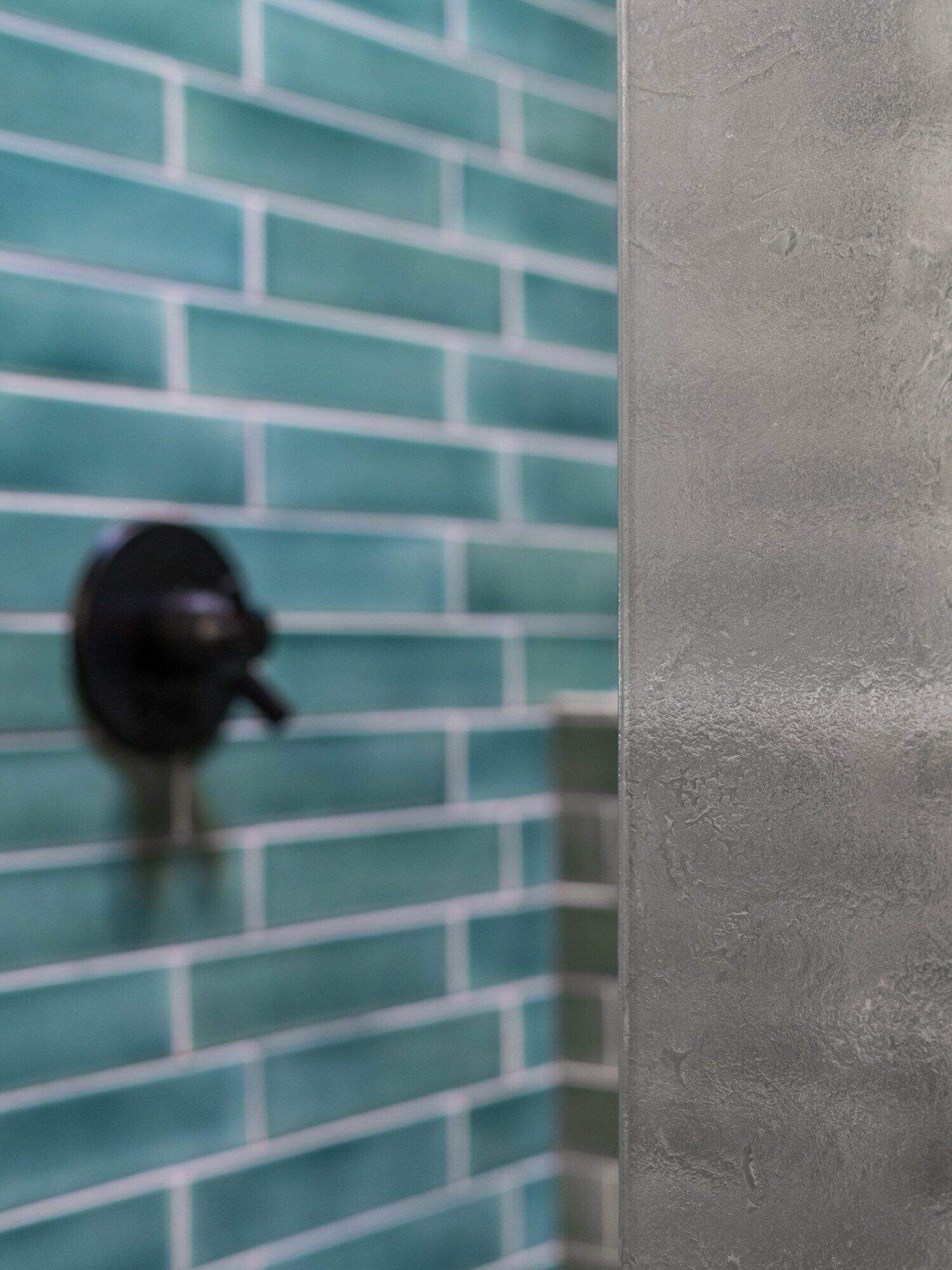 Close-up view of a shower with green subway tiles on the wall and a black faucet handle next to a frosted glass shower door.